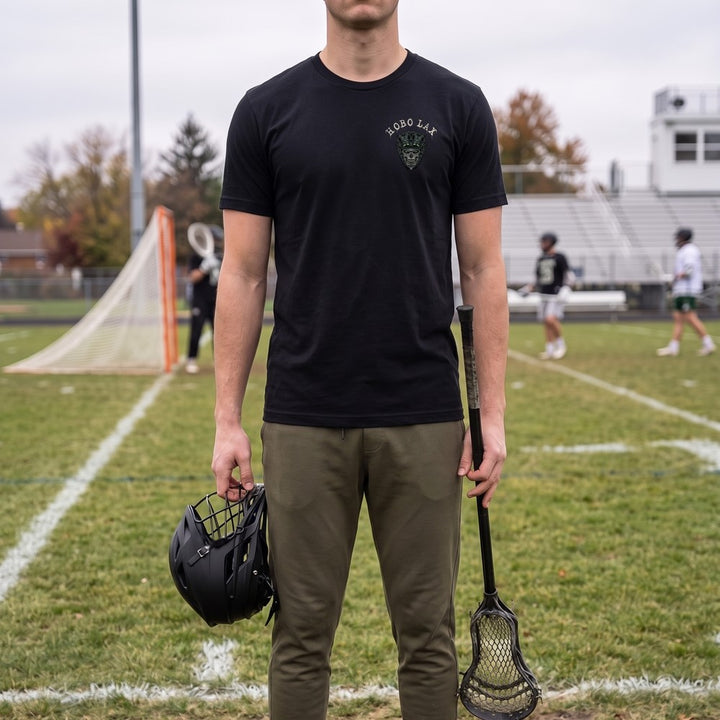 Man in the black Tactical Clear lacrosse tee with the small Hobo Lax skull logo on the left chest, holding his stick and helmet on the sideline
