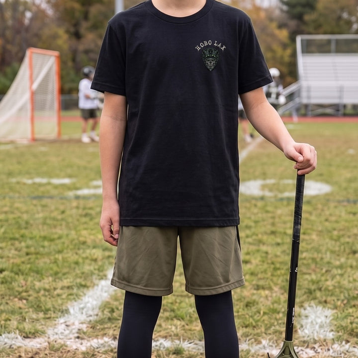 Boy in the black Youth Tactical Clear lacrosse tee with the Hobo Lax skull chest logo, holding his lacrosse stick on the sideline