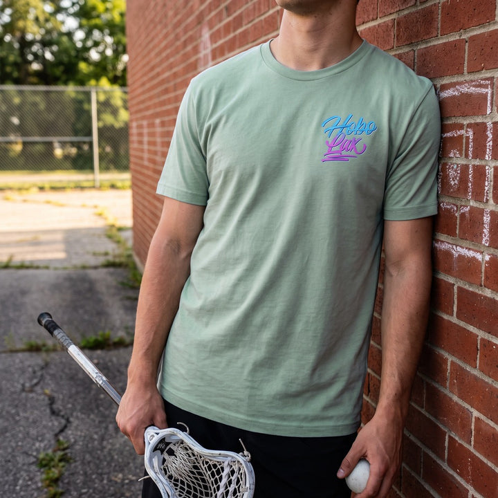Lax Snake Tee in seafoam leaning on a brick wall, front view with the Hobo Lax script logo on the chest