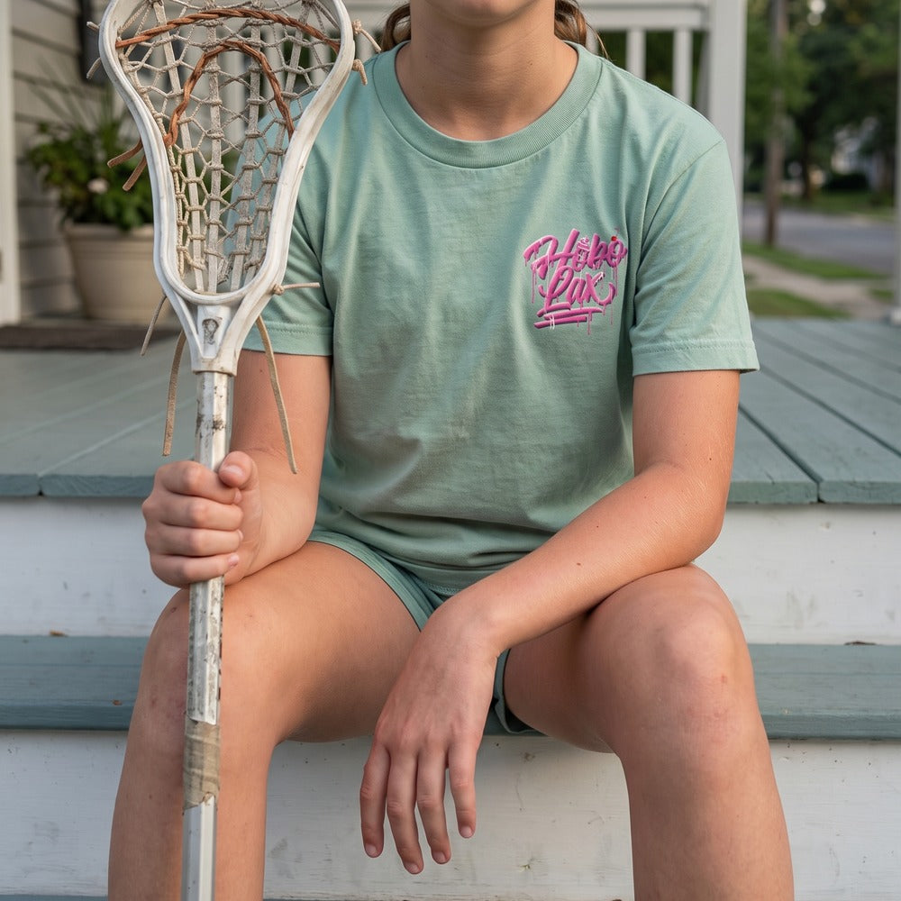 Youth Sideline Scoop Tee in seafoam worn by a girl sitting on porch steps, front view with the pink Hobo Lax drip logo on the chest